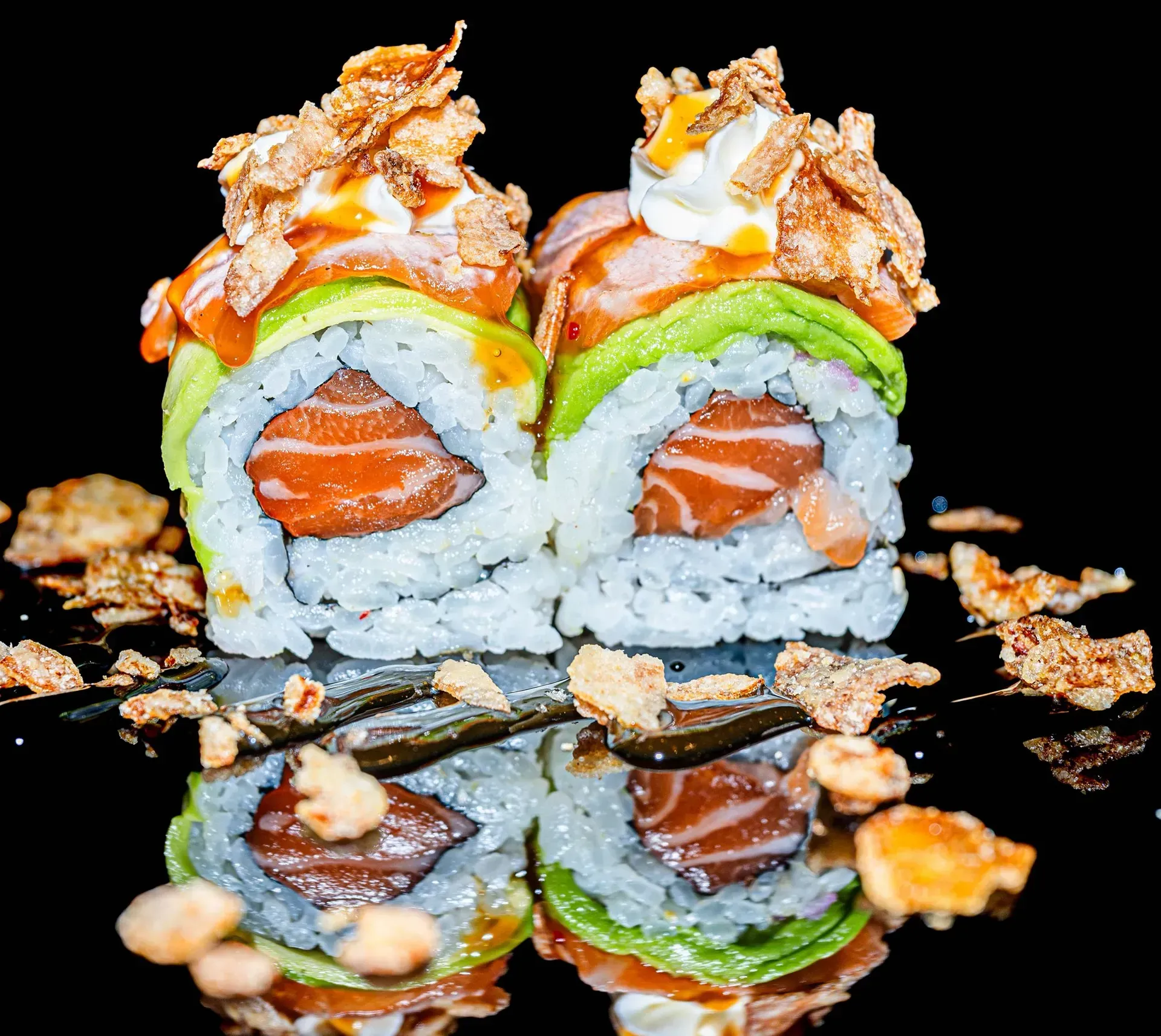 Metà di rotoli di sushi con salmone, avocado e guarnizione croccante su una superficie nera riflettente