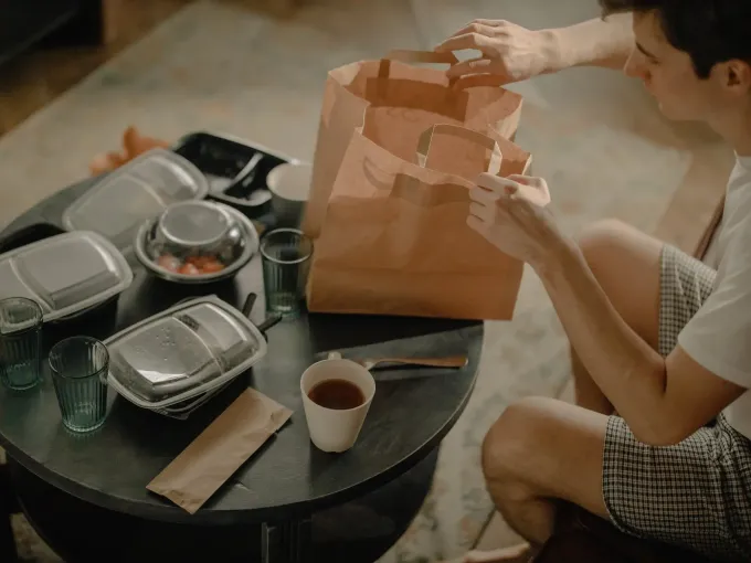 Una persona sta disimballando dei contenitori di cibo a un tavolo rotondo, accanto a un sacchetto di carta aperto e una tazza di caffè.