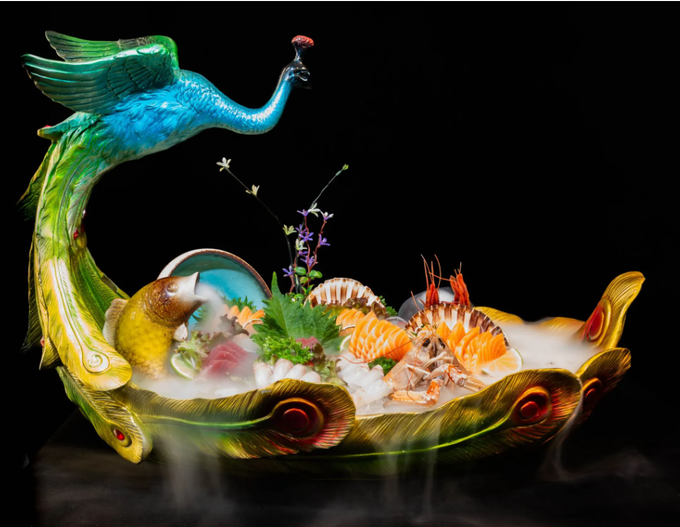 Un coloratissimo dessert fantastico a forma di pavone con frutta luminosa, fiori e dettagli arancioni su sfondo nero.