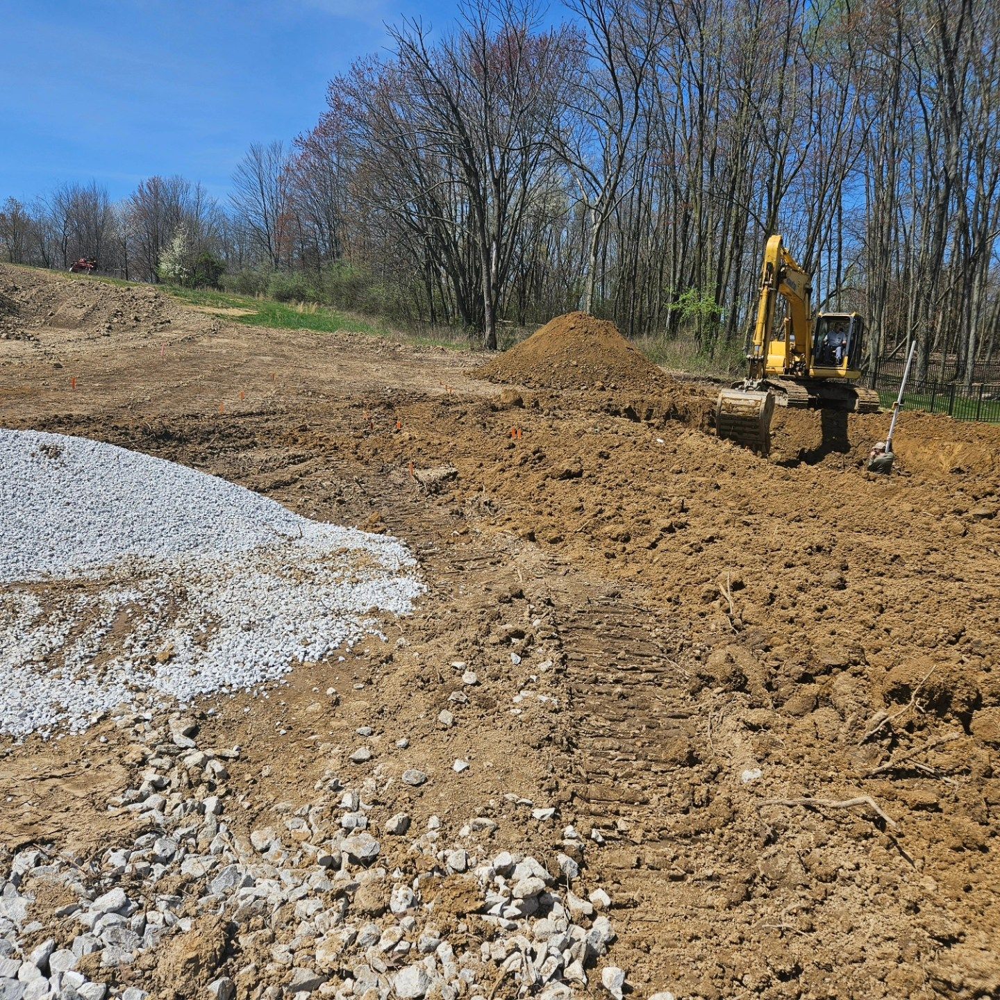 An excavator digs dirt near a pile of gravel in a construction site with trees in the background.