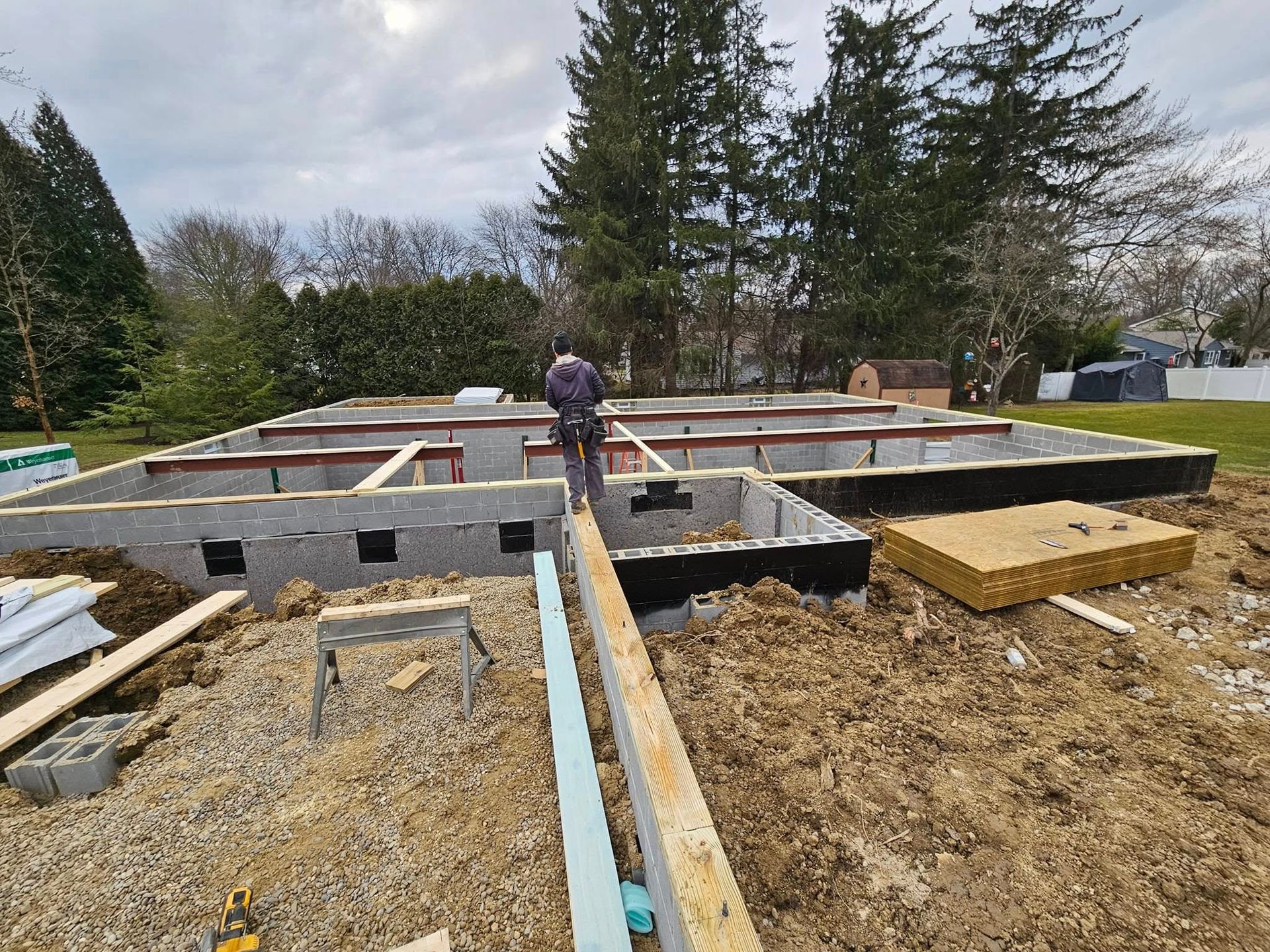Construction site, person working on a concrete foundation, building materials present, cloudy day.