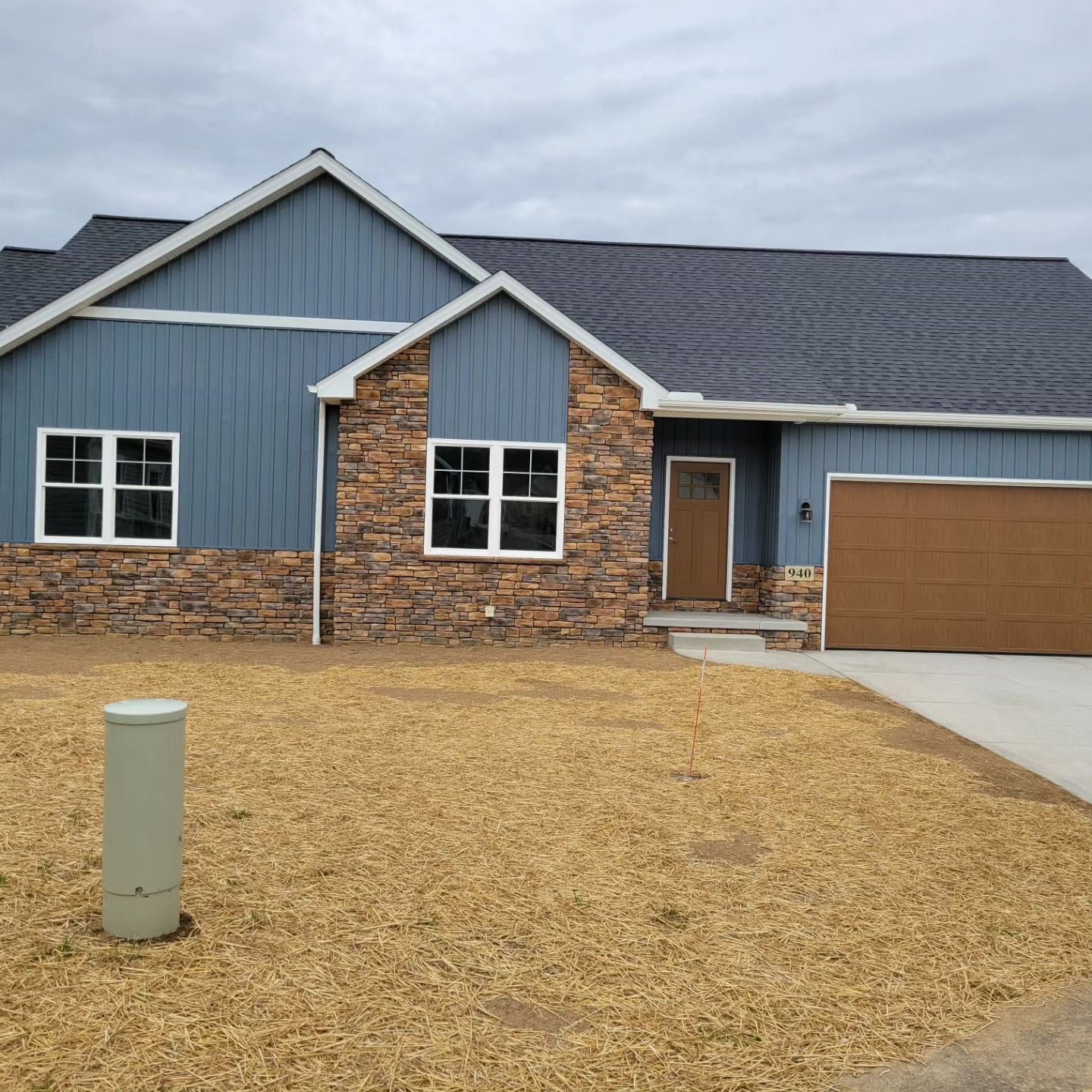 Blue and stone-colored house with a brown garage door on a gravel-covered lawn under a cloudy sky.