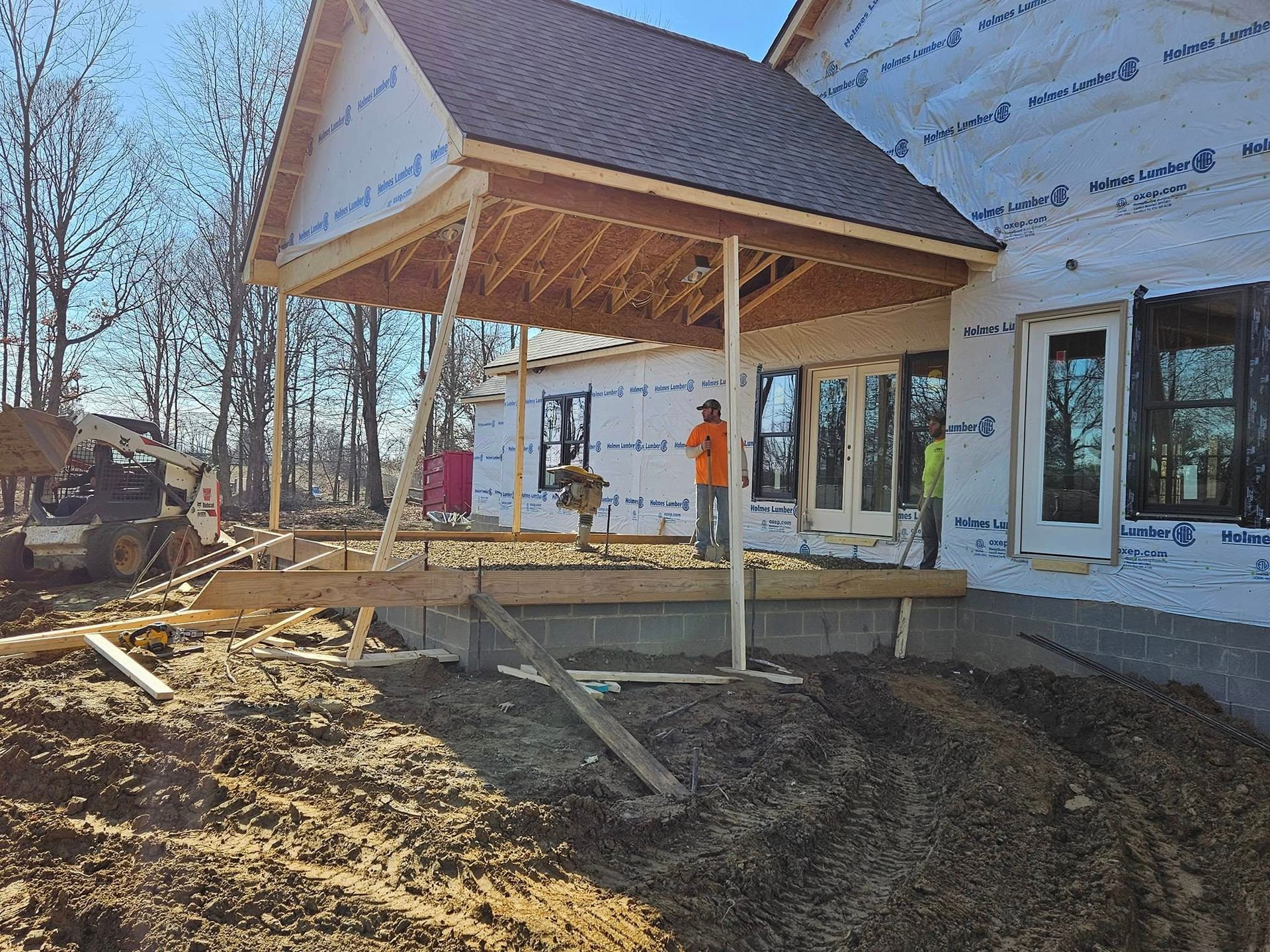 Construction site: porch roof framing over a concrete foundation. A worker stands nearby.