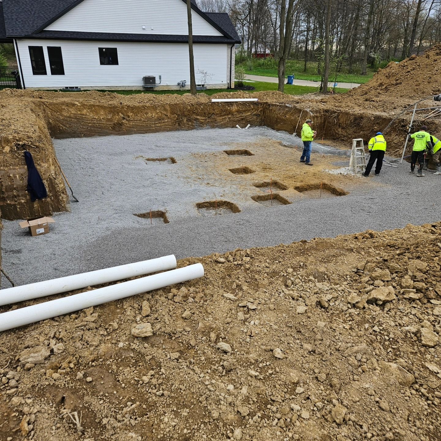 Construction site: excavated foundation with gravel, workers in yellow vests, and two white pipes.