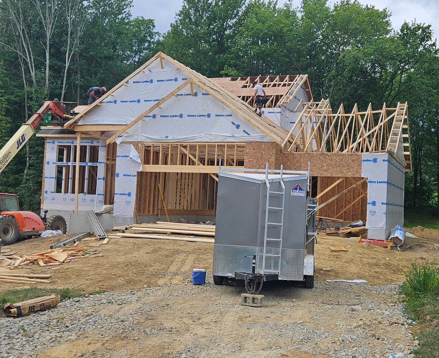 House under construction with exposed wooden frame and workers on the roof; trailer in front.
