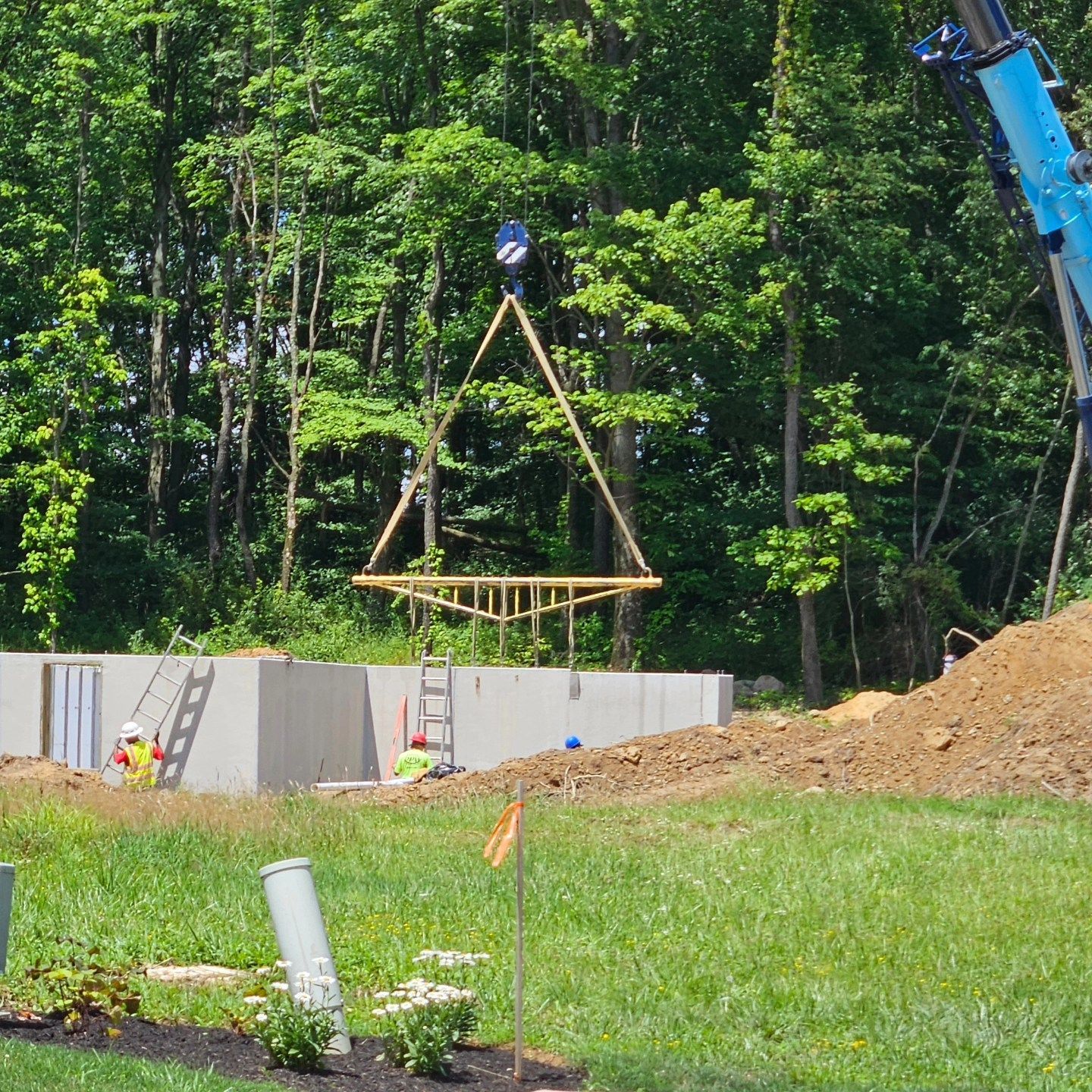 Construction site: A triangular roof frame is lifted by a crane over a concrete foundation. Workers in hard hats.