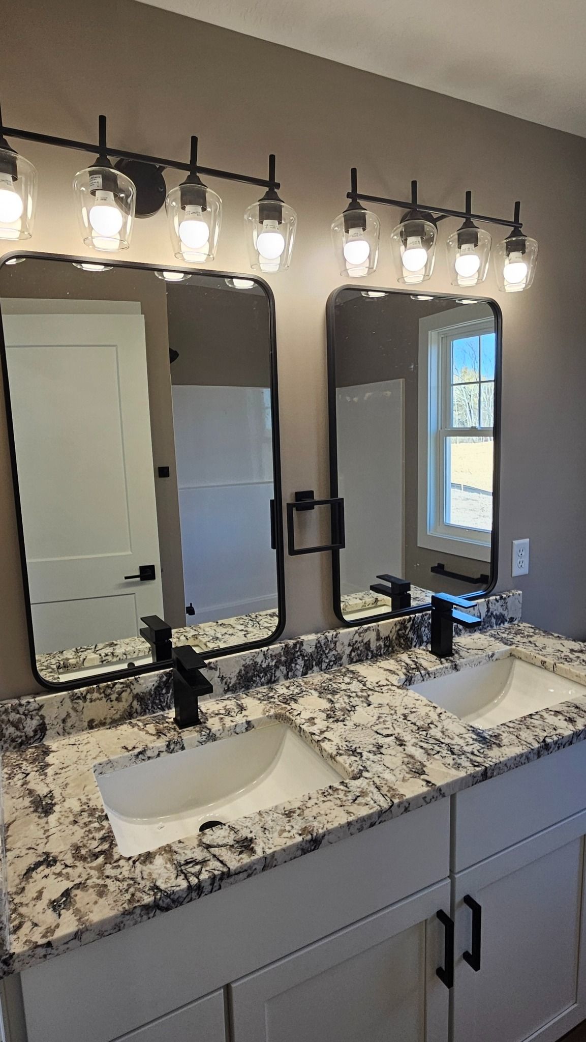 Bathroom vanity with dual sinks, mirrors, black faucets, and lights. Granite countertop, white cabinets, beige walls.