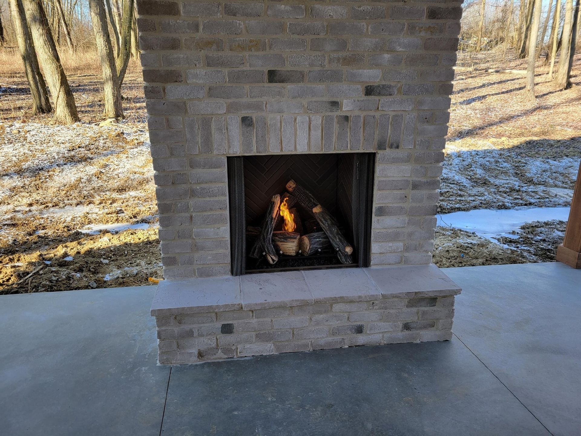 Outdoor stone fireplace with a fire burning, set on a patio. Trees in the background.