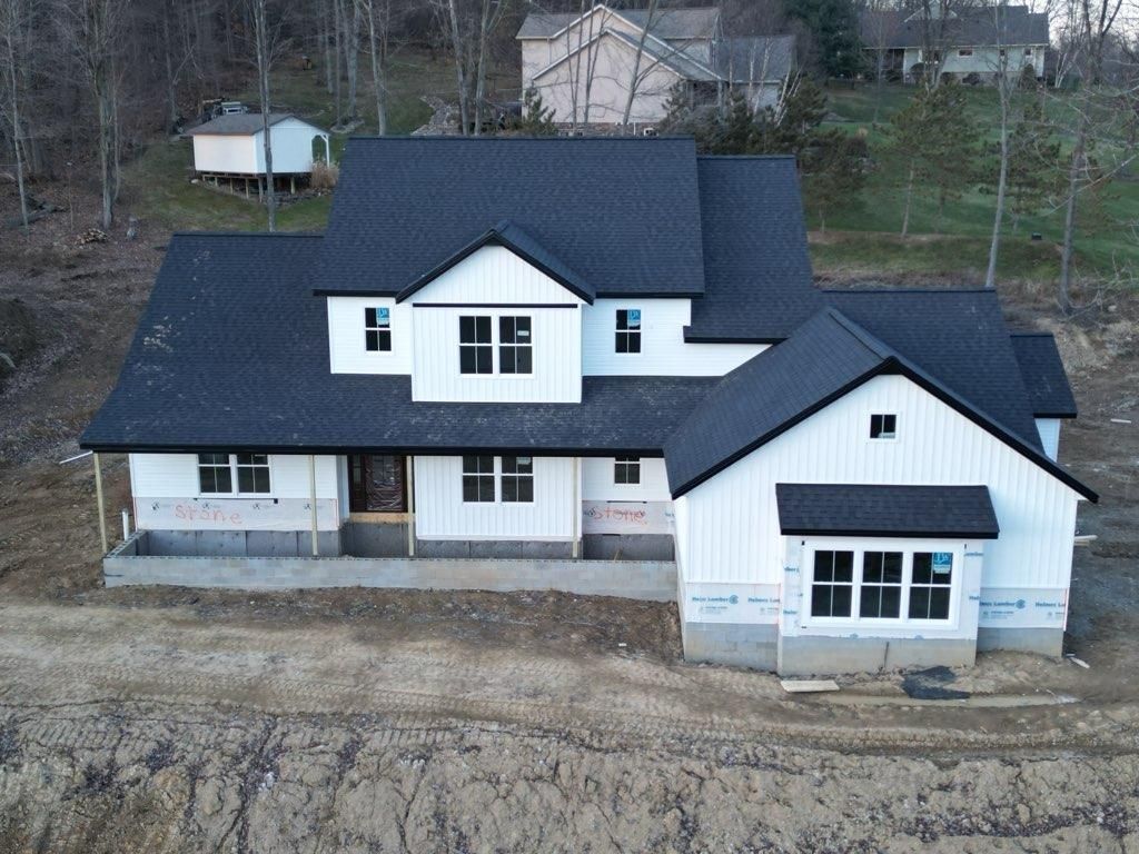 New white farmhouse-style house with black roof under construction on a hillside.