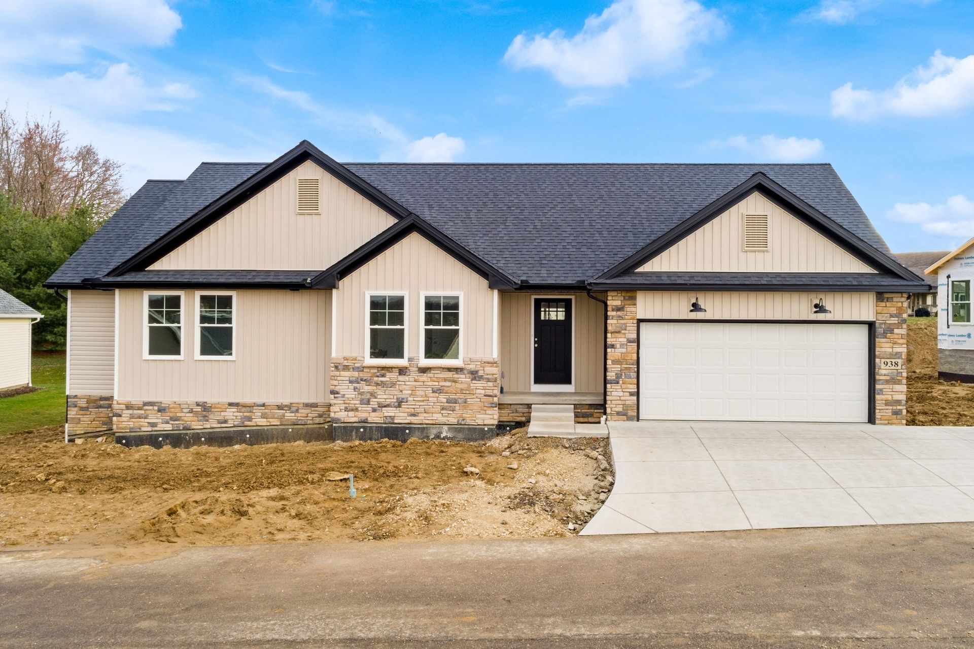 Tan house with black roof and stone accents, concrete driveway, overcast sky.