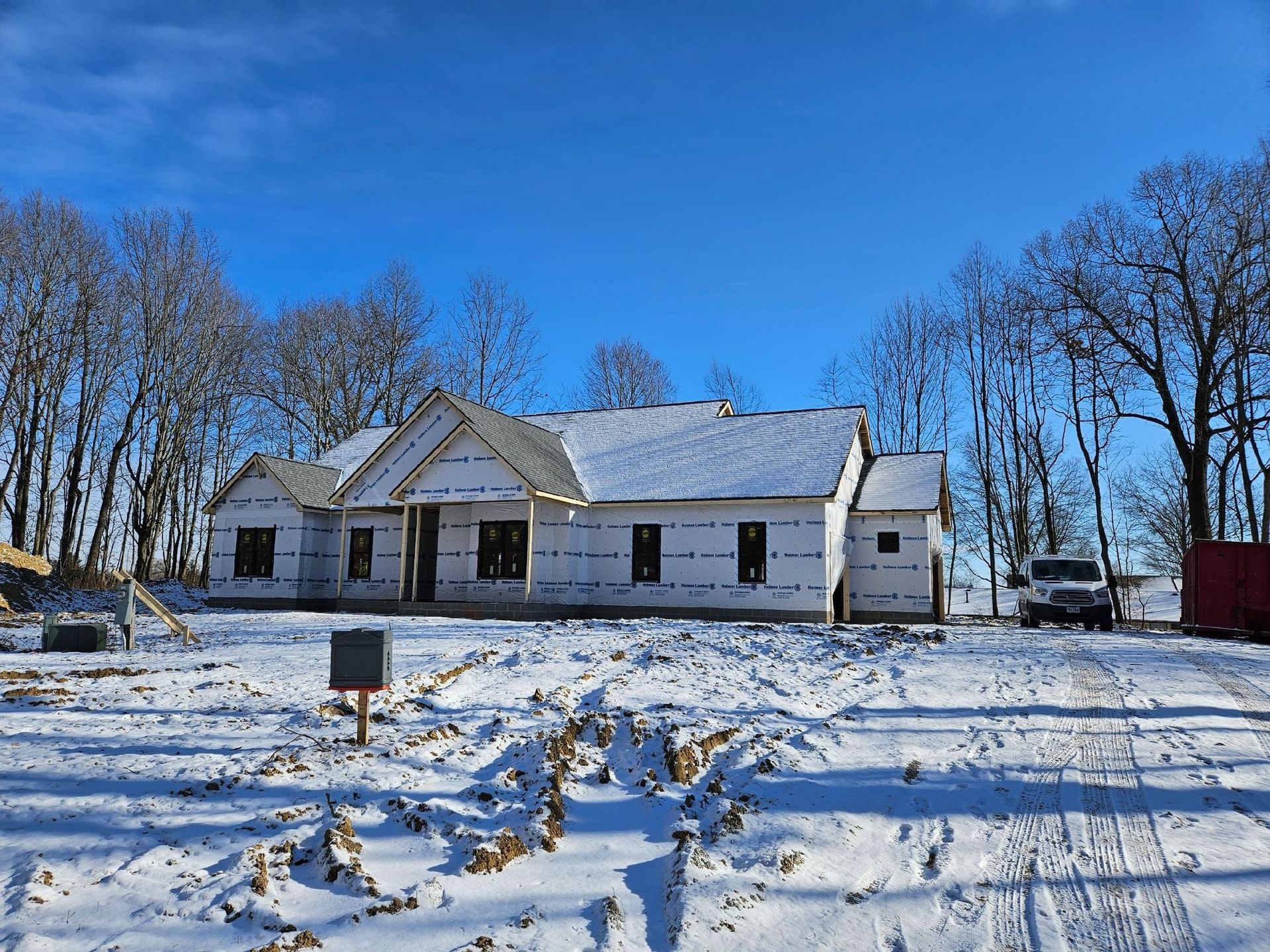 New house under construction, covered in white wrap, in a snowy landscape on a sunny day.