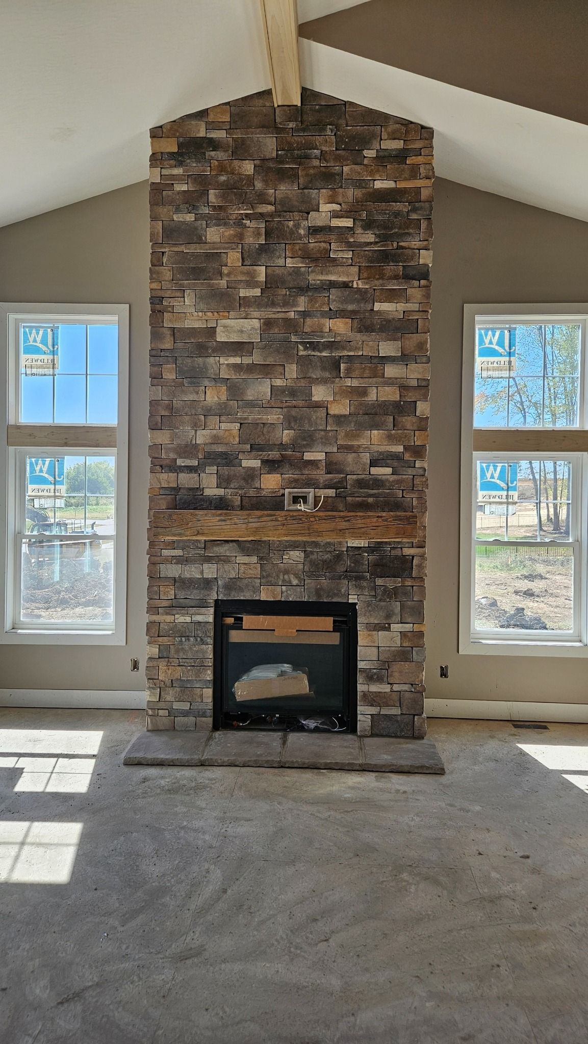 Fireplace with stone facade between two windows in a room with a concrete floor and a vaulted ceiling.