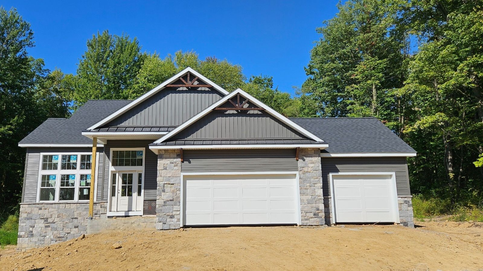 New house with grey siding, stone accents, and two-car garage. Overcast sky and surrounding trees.