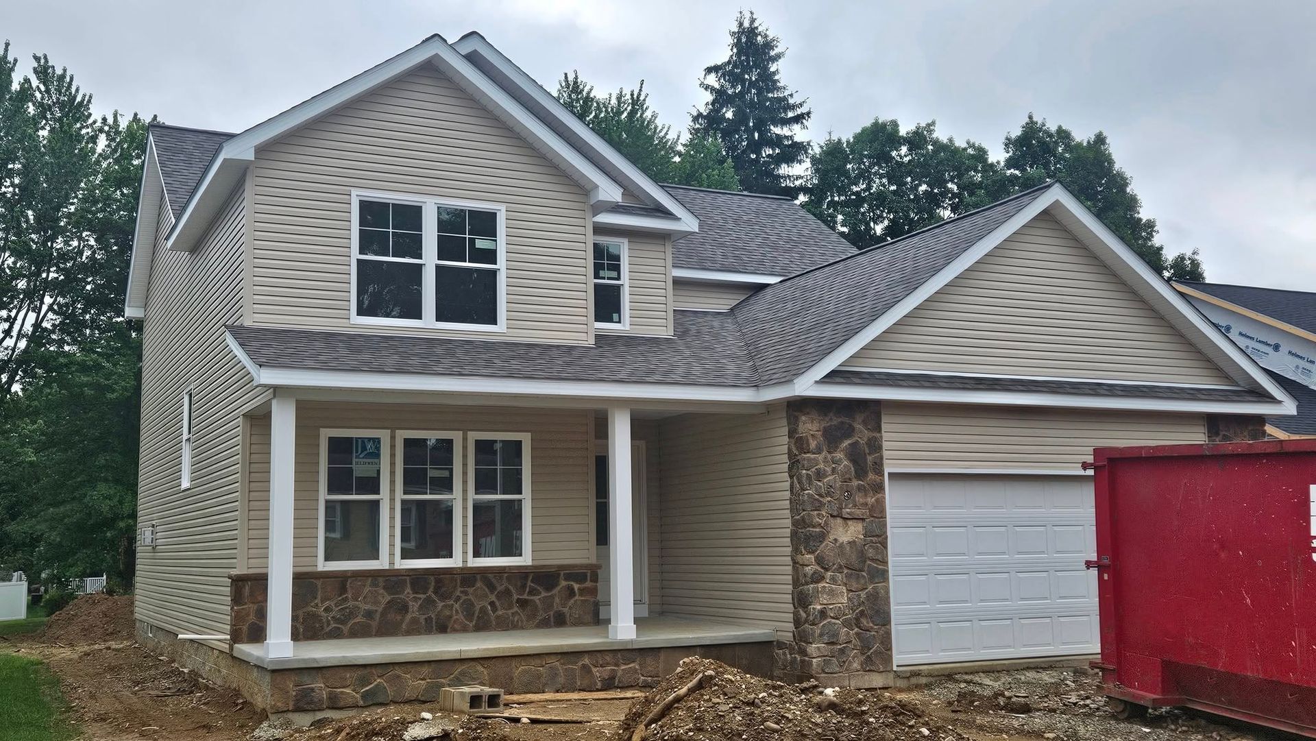 New two-story house under construction, with tan siding, stone accents, and a red dumpster in the yard.