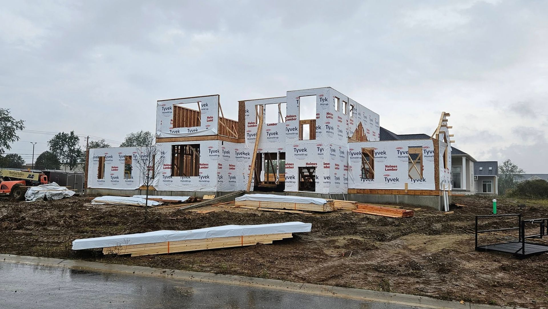 House under construction, wood framing and sheathing visible on a muddy lot under an overcast sky.