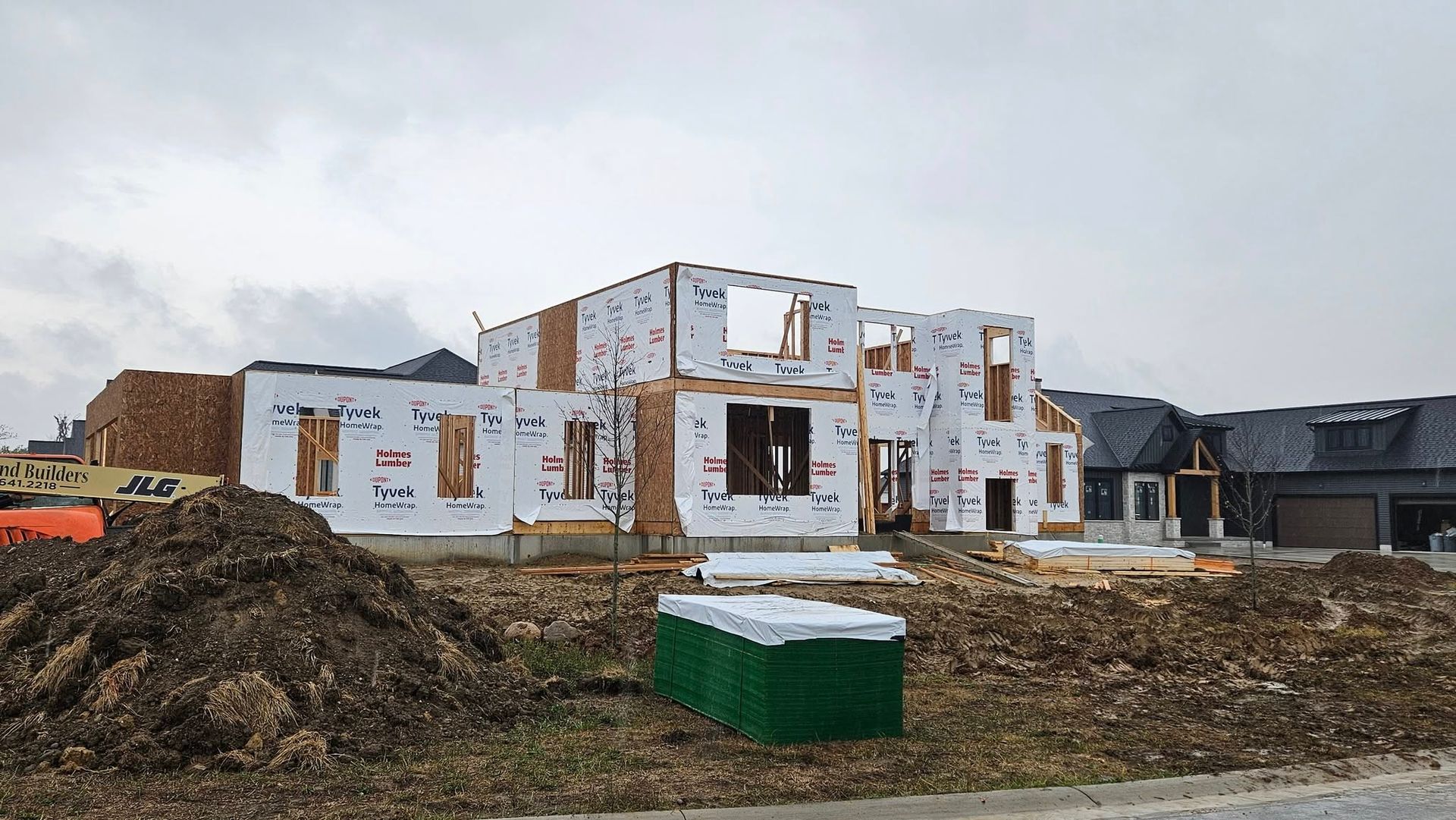 A house under construction; wood frame and sheathing visible. Earth mound in the foreground. Overcast sky.