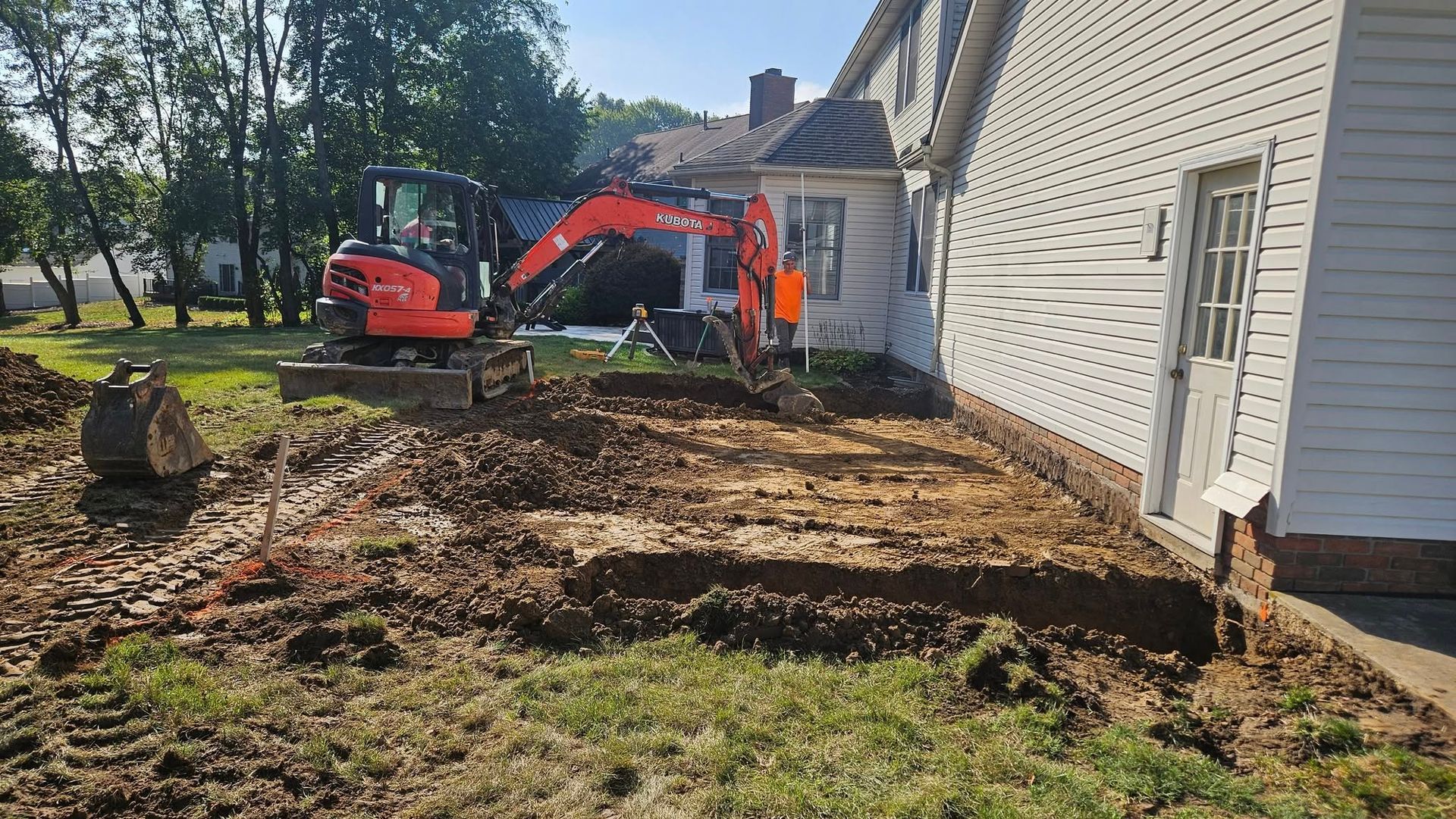 An excavator digs near a house, creating a trench. Green grass and blue sky.