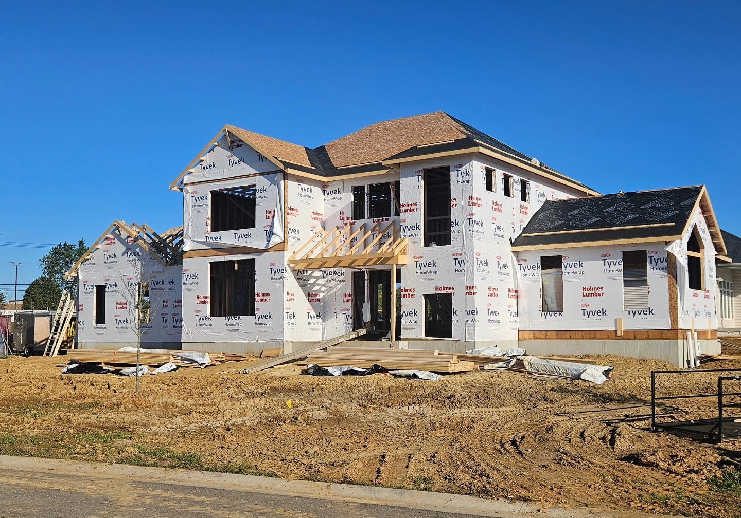 Two-story house under construction, wrapped in weather-resistant barrier, with dark window frames and blue sky.