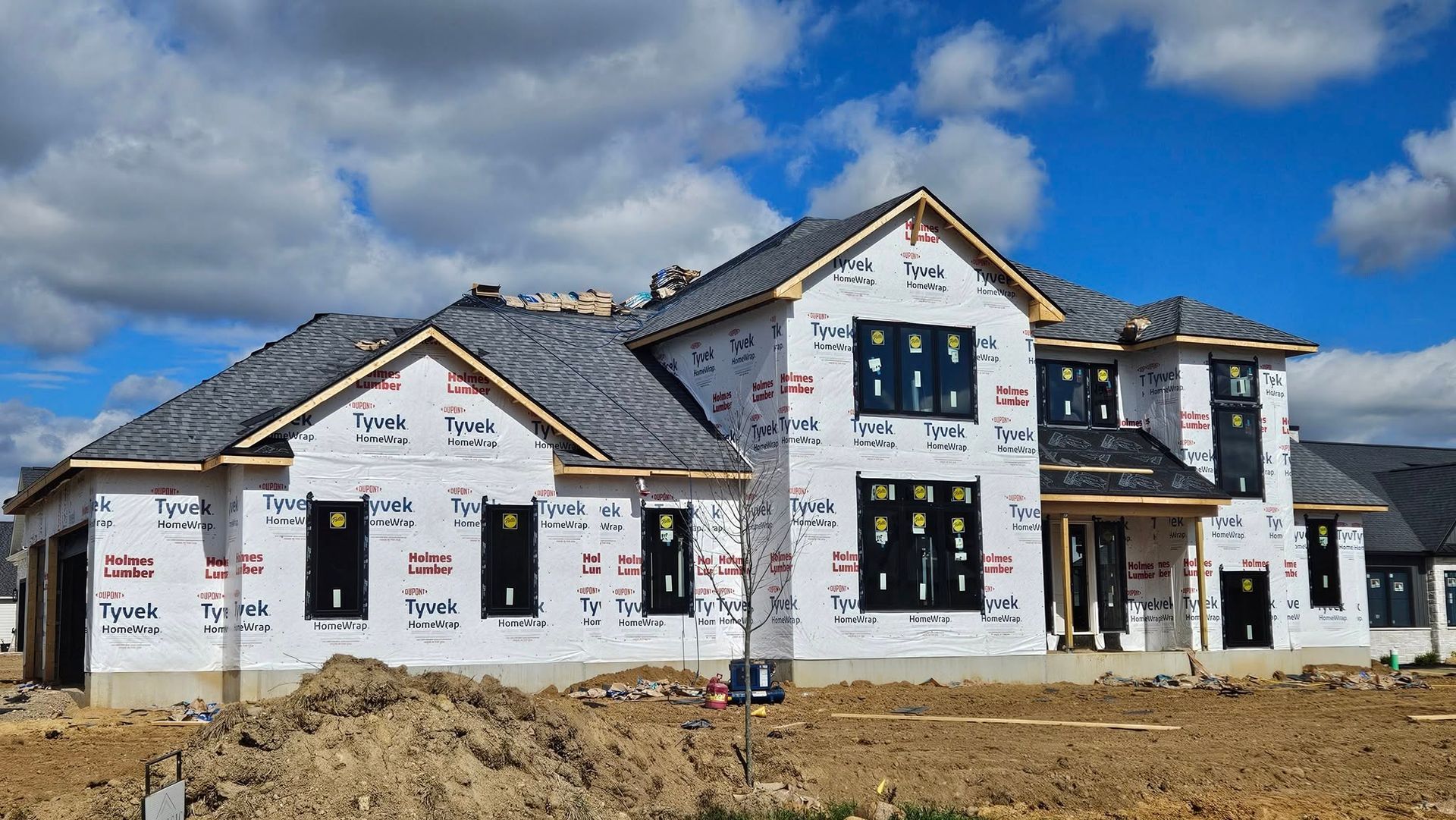 Two-story house under construction, wrapped in white material, with dark window frames and a gray roof, against a cloudy blue sky.
