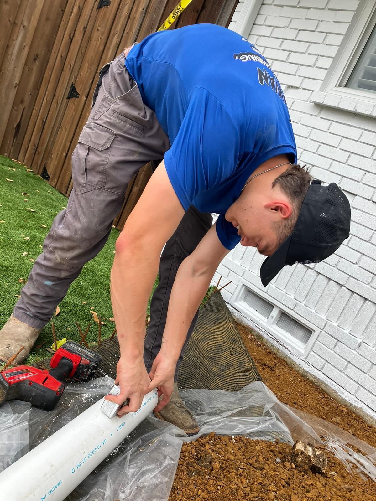 A man in a blue shirt is working on a pipe outside of a house.