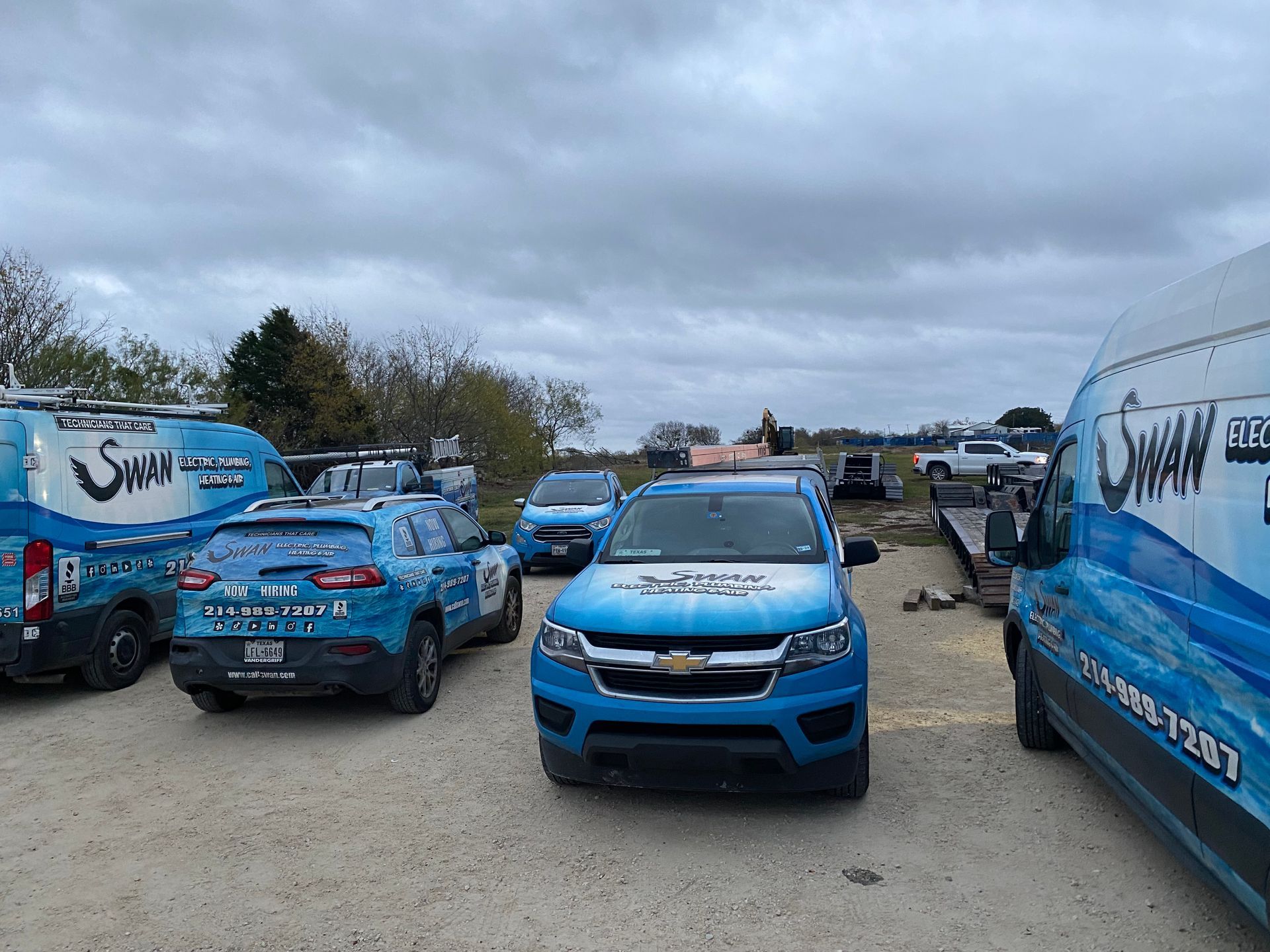A group of blue trucks and vans are parked in a gravel lot.