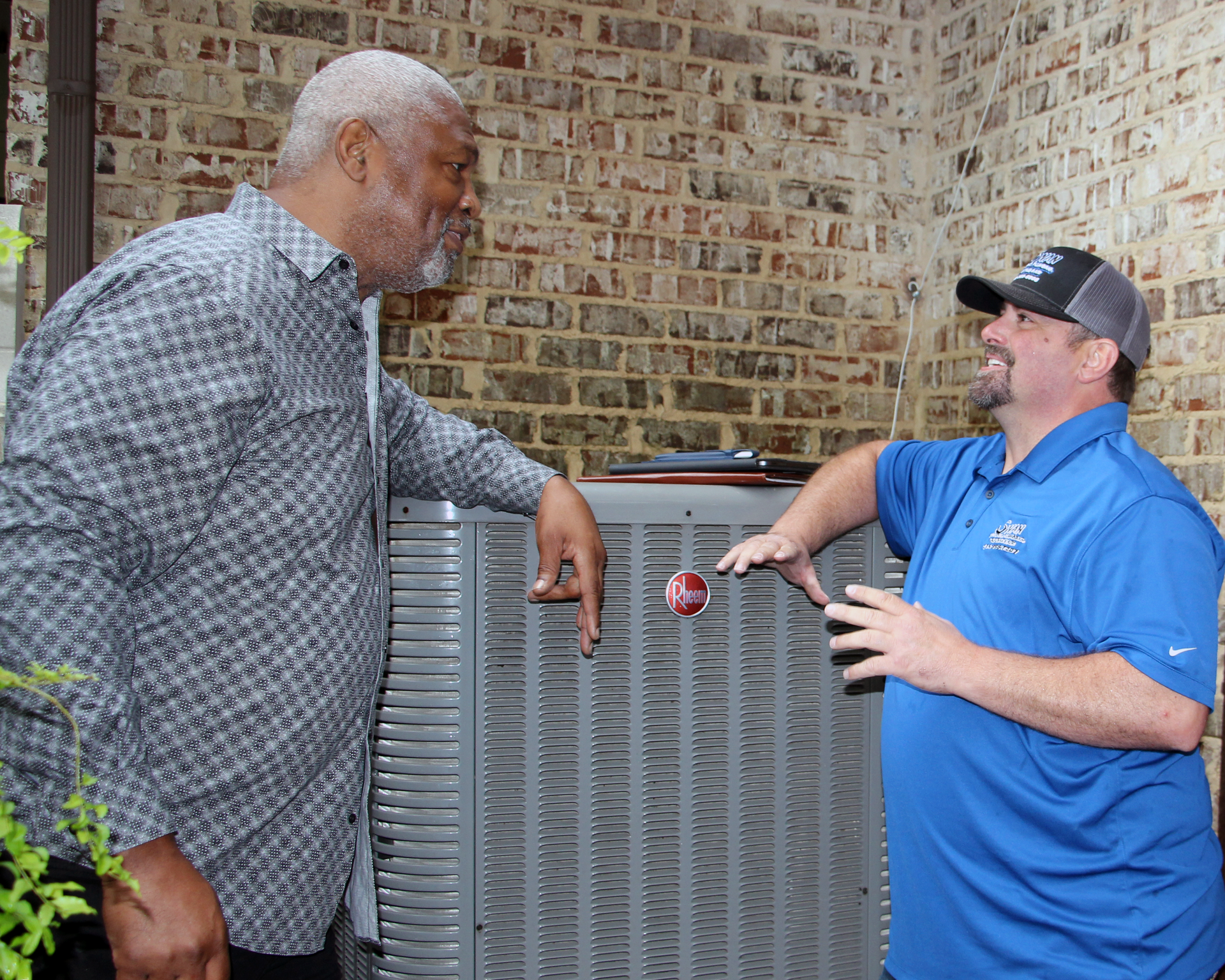 Two men are standing next to each other in front of an air conditioner.