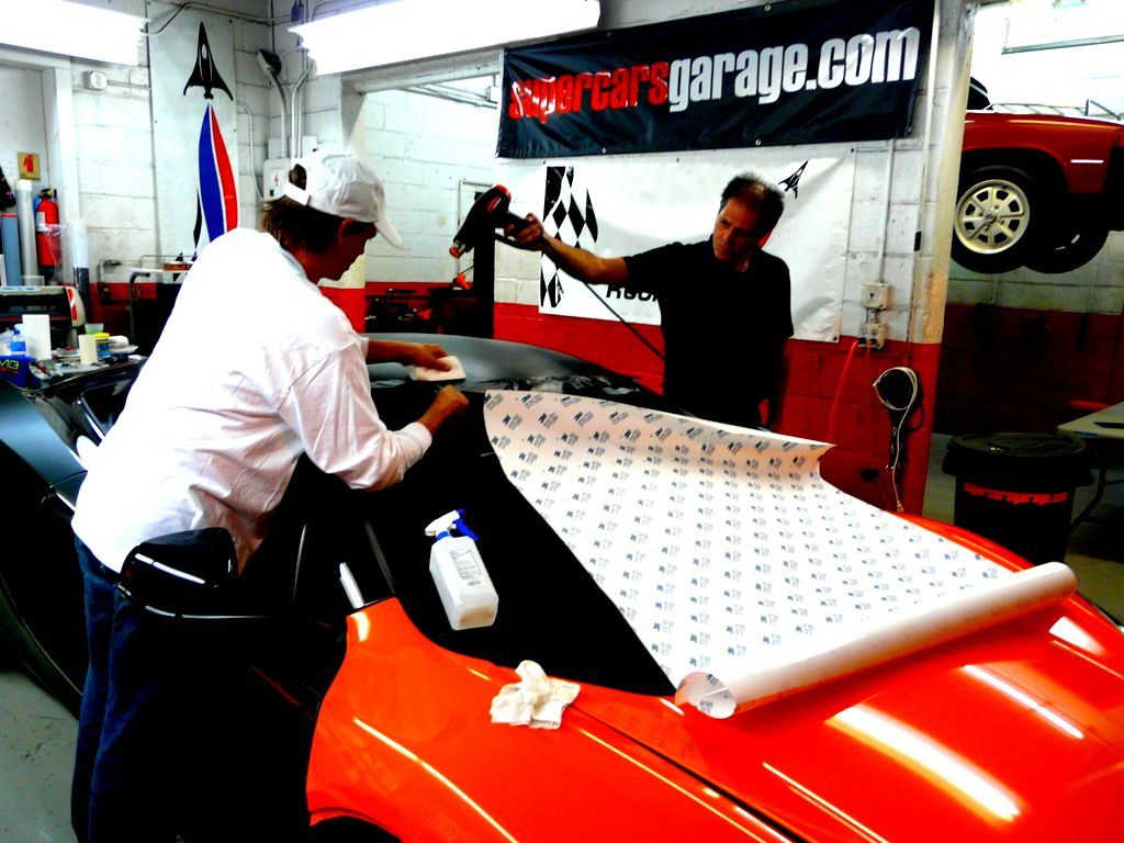 Two people applying a wrap to a car in a garage; one uses a heat gun.