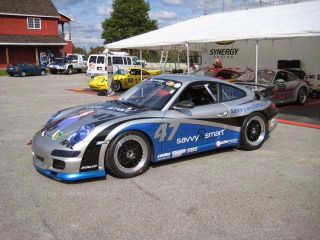 Silver and blue race car, number 47, parked near a white tent. Other vehicles and a building are in the background.
