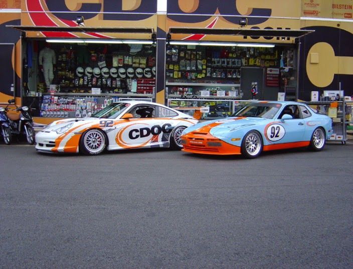 Two sports cars parked outside a shop: white and orange Porsche, blue and orange Porsche 944.