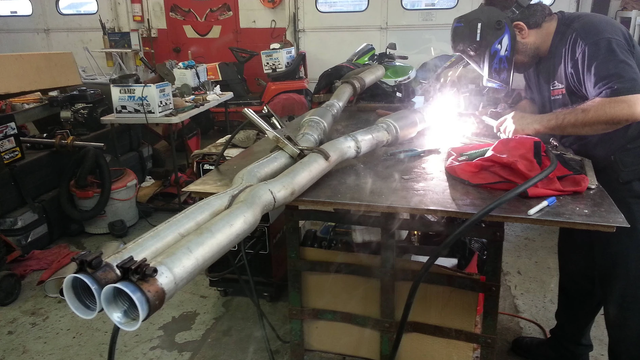 Man welding exhaust system on workbench in a garage.