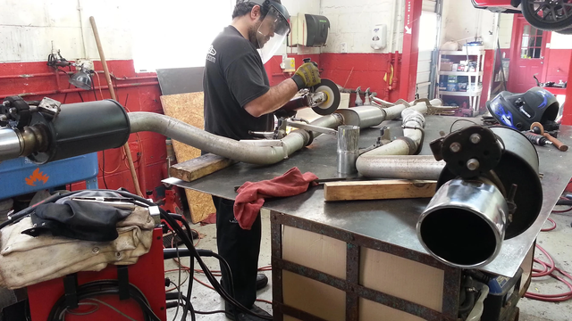 A mechanic welding car exhaust pipes in a garage. Tools and car parts surround him.