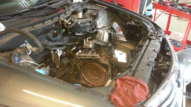 Car engine bay with components exposed; a mechanic is working on it.