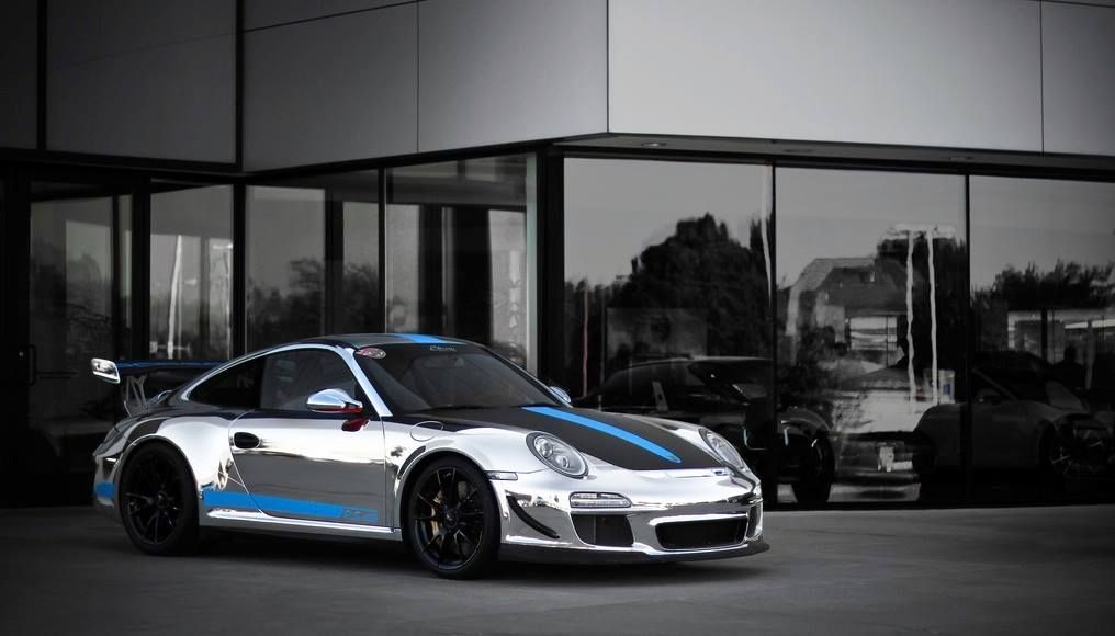 Chrome-finished Porsche sports car with blue racing stripes parked in front of a modern building with reflective windows.