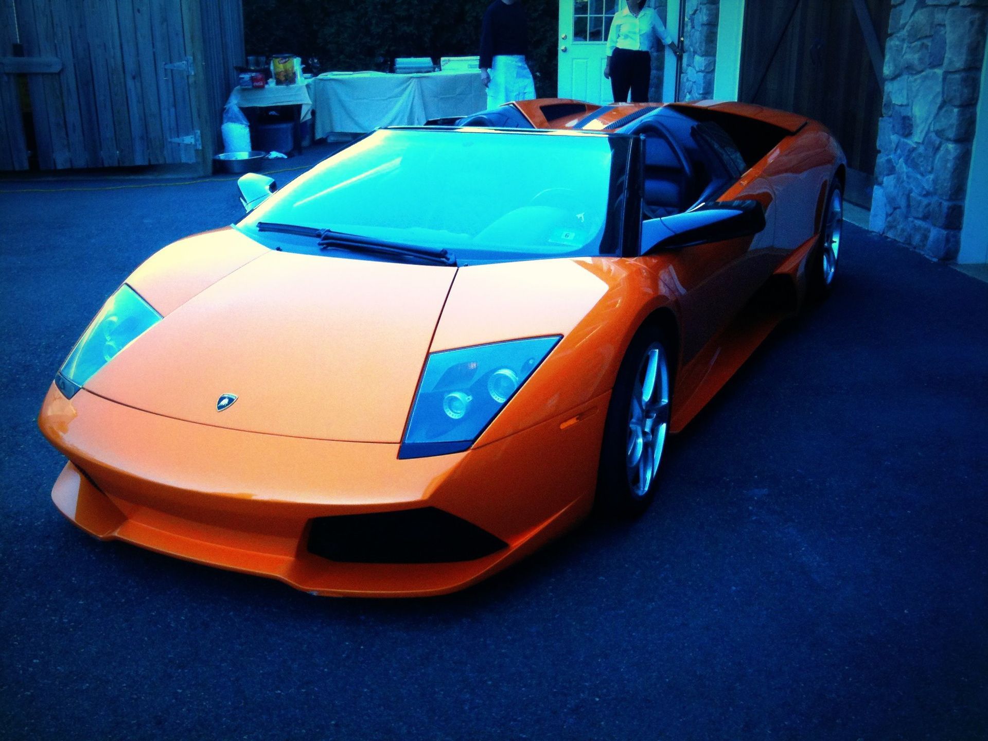 Orange Lamborghini parked on a blue-lit driveway; a person is visible in the background.