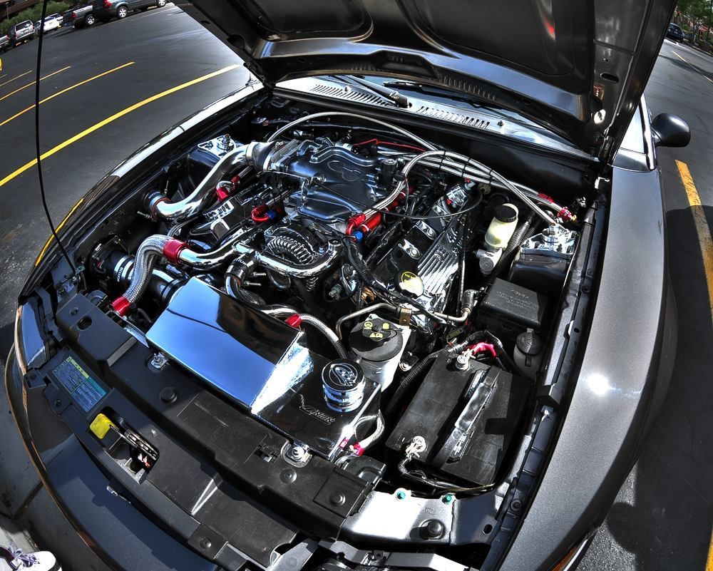 Black car engine bay with chrome and red accents; hood open in a parking lot.