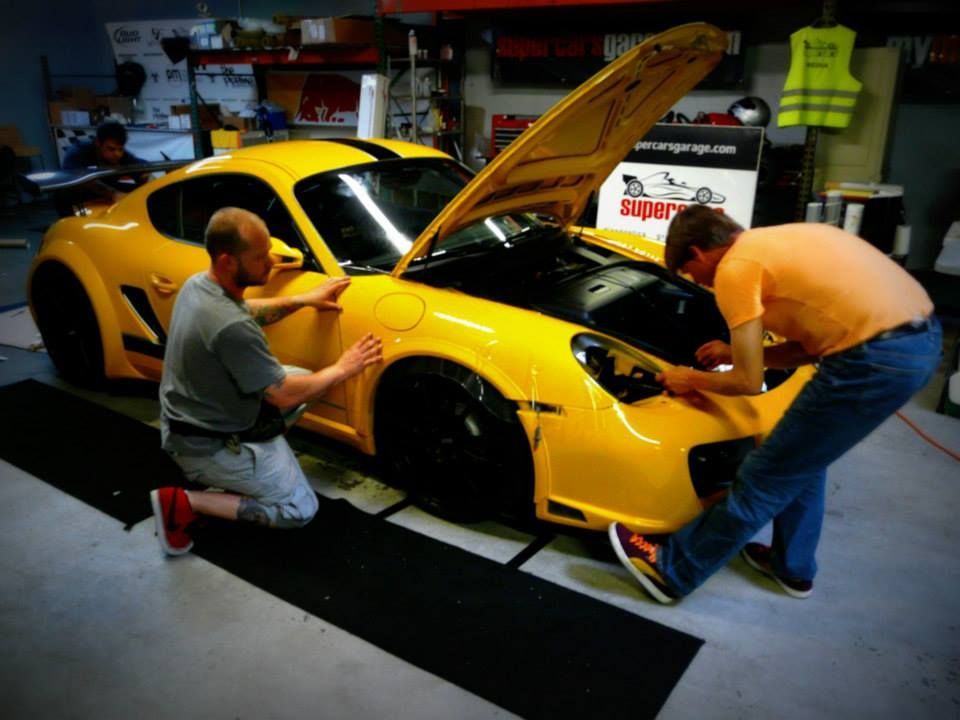 Two men working on a yellow Porsche with the hood up in a garage.