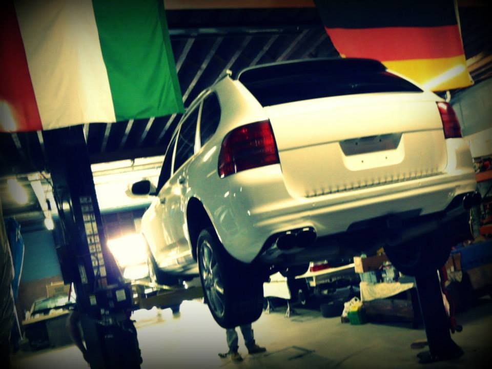White Porsche SUV on a lift in a garage, with Italian and German flags hanging above.