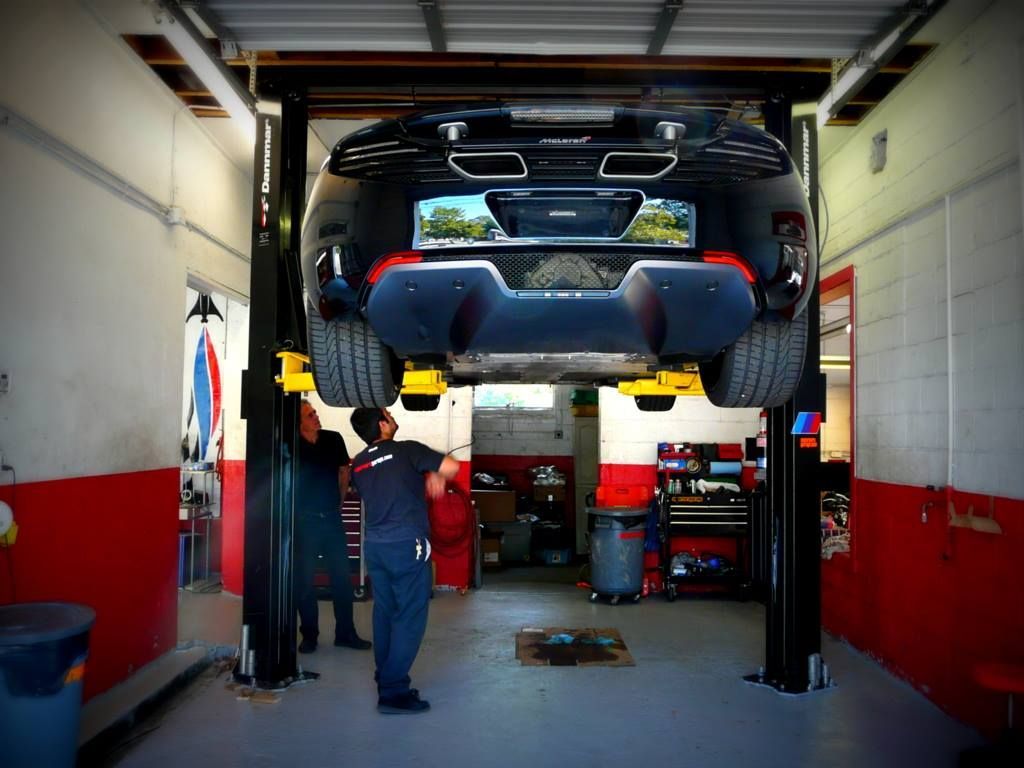 A black car raised on a lift in a mechanic shop; two mechanics work underneath it.
