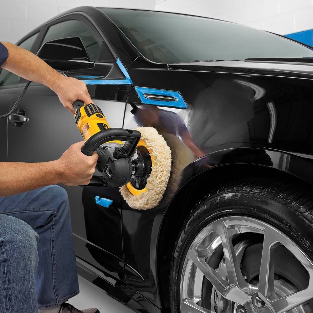 Man polishing black car with a rotary buffer in a garage. Blue tape protects some areas.