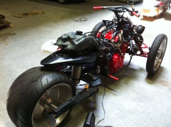 Custom three-wheeled motorcycle with a large rear tire, black body, and exposed engine. Garage setting.
