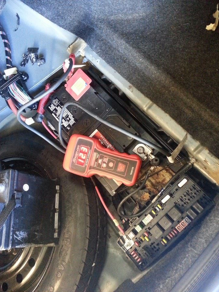 Car battery, fuse box, and a red multimeter in the trunk of a vehicle.