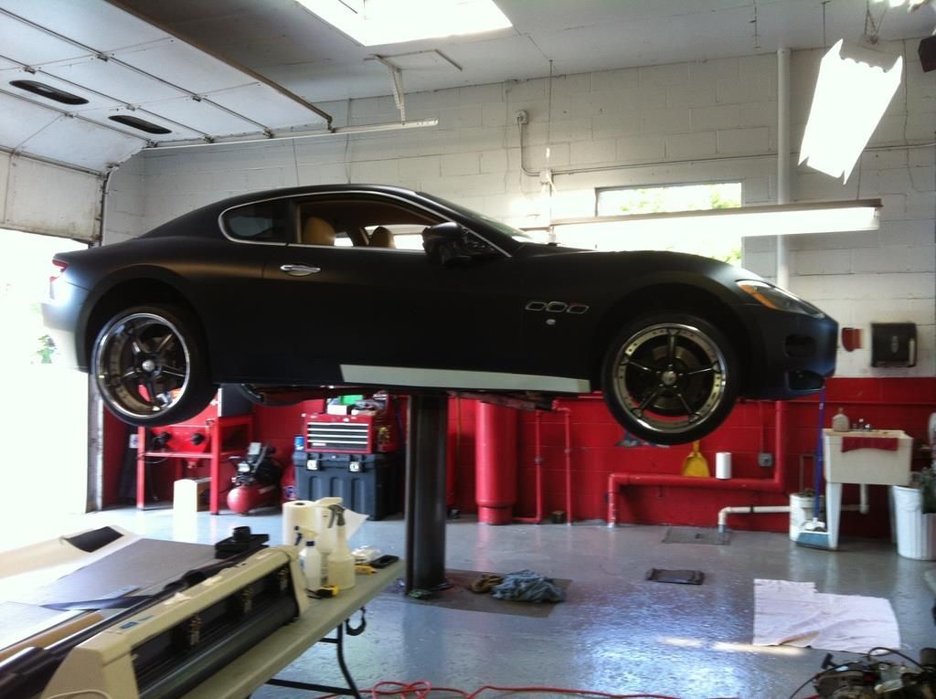Black sports car on a lift in a garage. Red walls, grey floor, and white equipment visible.