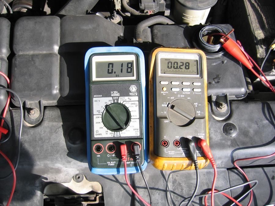 Two multimeters on a car engine, red and black probes connected. Blue multimeter reads 0.18, yellow reads 0.028.