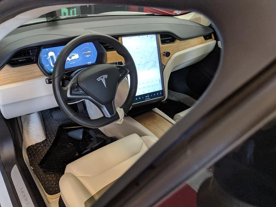 Tesla car interior: steering wheel, large central display, cream-colored seats, wood trim, and floor mat.