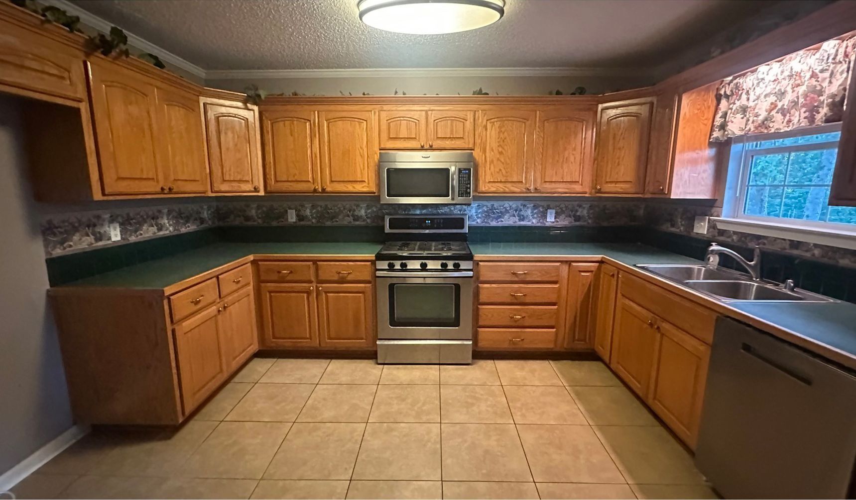 U-shaped kitchen with light brown cabinets, stainless steel appliances, green countertops, and tan tile flooring.