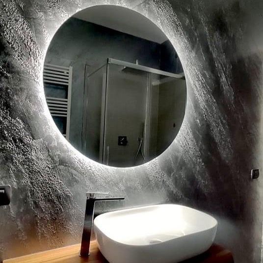 In un bagno moderno, uno specchio rotondo retroilluminato è appeso sopra un lavabo bianco a ciotola, su una parete grigia rustica e texturizzata.