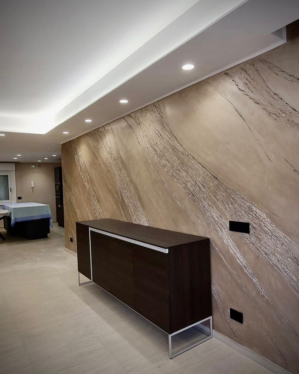 Una credenza elegante color marrone scuro è addossata a una parete con una finitura in gesso decorativo beige, caratterizzata da una texture diagonale.