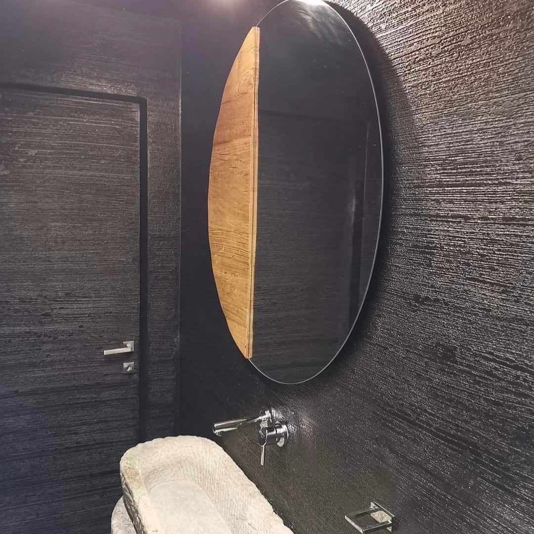 Un bagno moderno con pareti scure e texturizzate, un lavabo in pietra, un rubinetto argentato e uno specchio rotondo con un inserto laterale in legno.