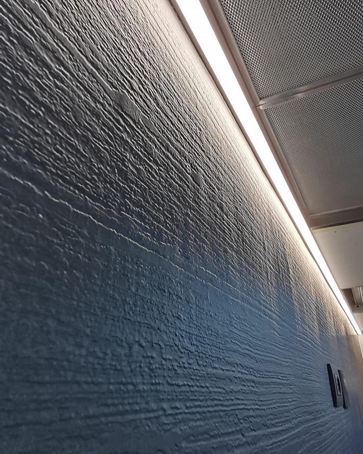 Vista dal basso di una parete grigia con superficie ruvida, illuminata da una luminosa striscia LED orizzontale incassata nel soffitto.