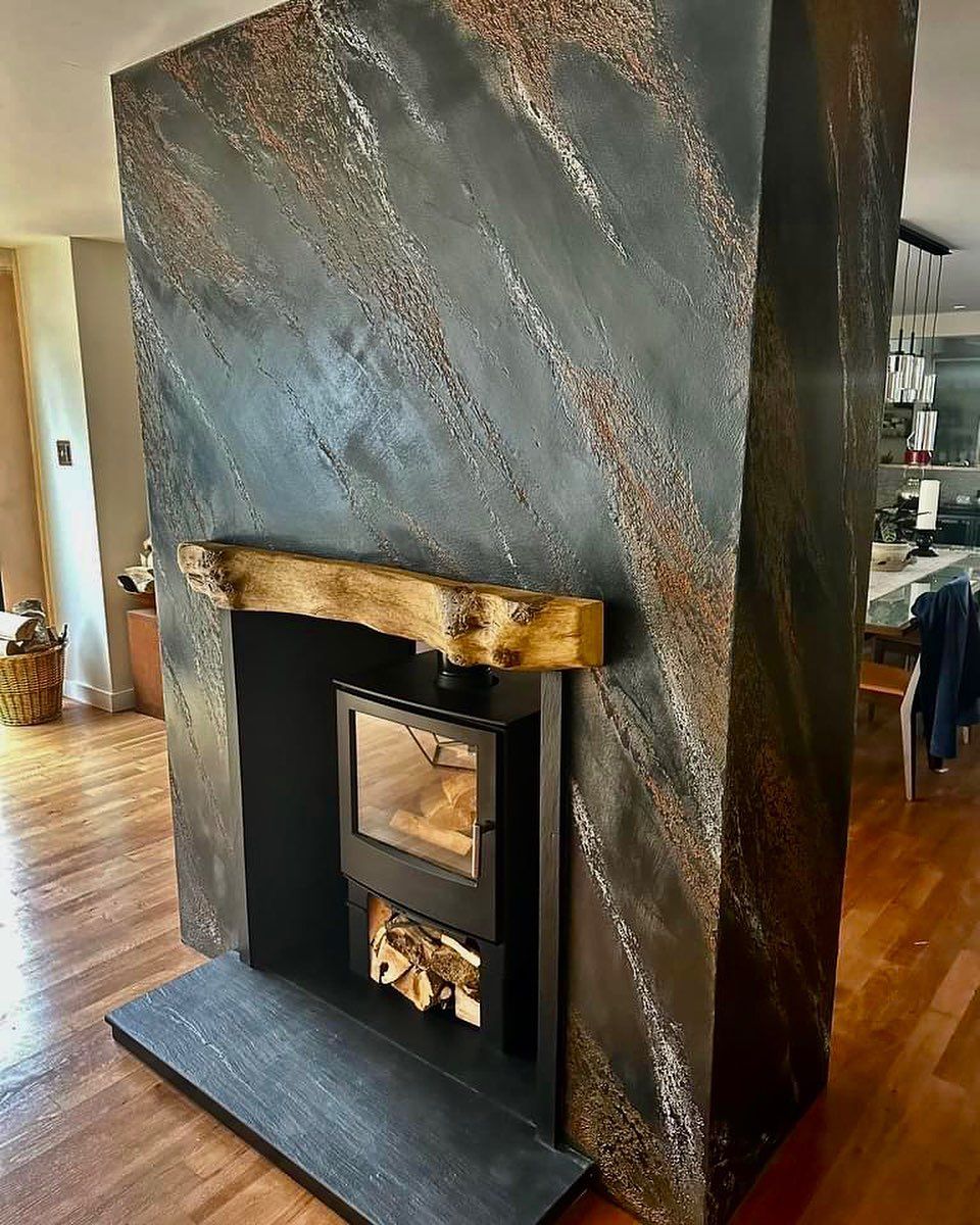 Un camino moderno caratterizzato da una finitura scura e materica color antracite e rame, con una mensola in legno rustico e una stufa a legna.